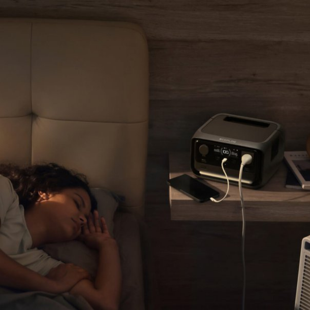 Person sleeping on a couch next to a power station and phone in a dimly lit room.