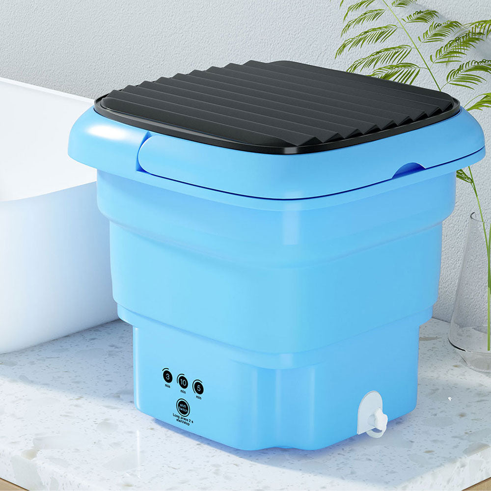 Blue collapsible washing machine with black lid placed next to a plant on tiled floor.