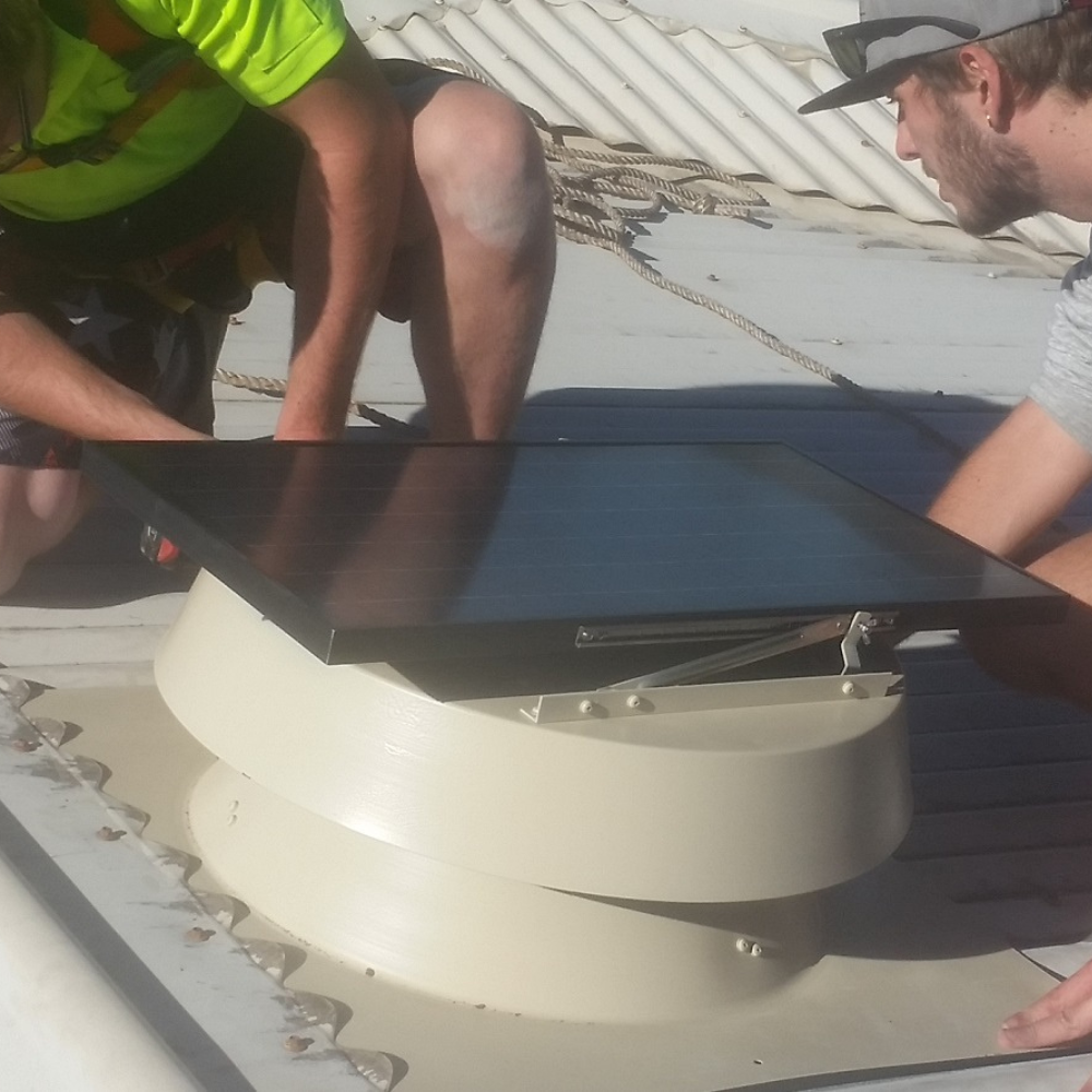 Two workers installing a silver solar roof ventilator on a corrugated metal roof.