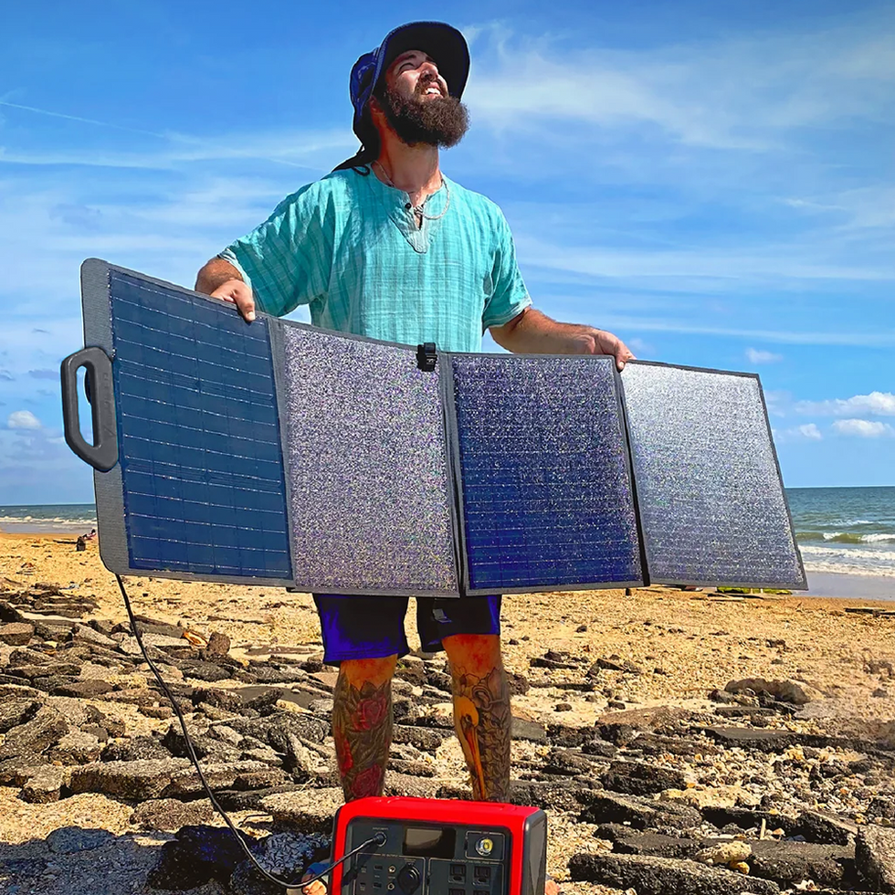 A sunny beach setting with a person holding a portable solar panel system, standing on the sandy shore. A portable power station is placed at their feet, symbolizing energy independence and renewable power in remote locations.