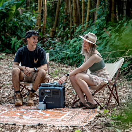 Couple sitting in a forest clearing with Bluetti power station between them, illustrating off-grid power during outdoor camping.