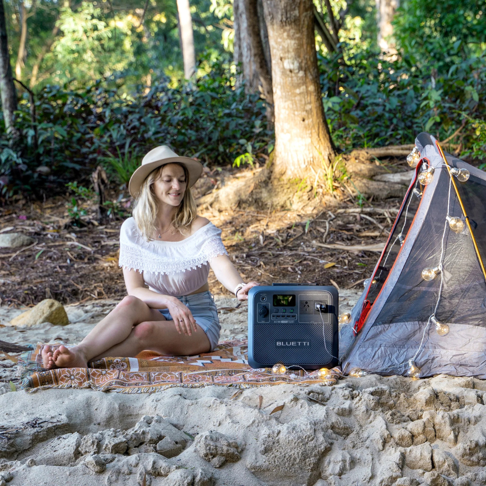 Woman reading beside a lit campfire with Bluetti unit nearby, showcasing peaceful night-time use in a wooded setting.