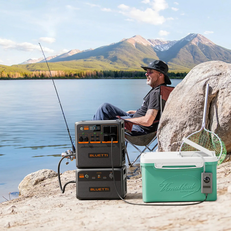Man fishing by a lake with Bluetti power station nearby, powering a portable fridge, illustrating outdoor recreational use.