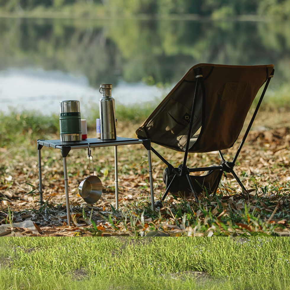 Outdoor camping setup by a serene lakeside with a brown folding chair and a small table holding a thermos, water bottle, and camping gear