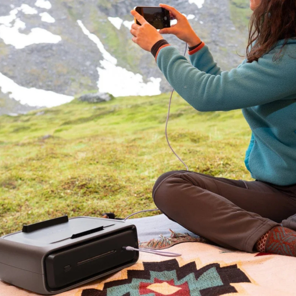 Person sitting outdoors using a portable device, with mountains and grass in the background