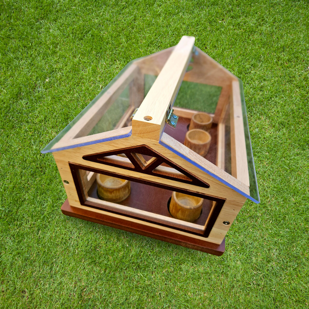 Angled View on Grass
A mini wooden and glass greenhouse with the roof slightly opened, sitting on green grass in an outdoor setting.