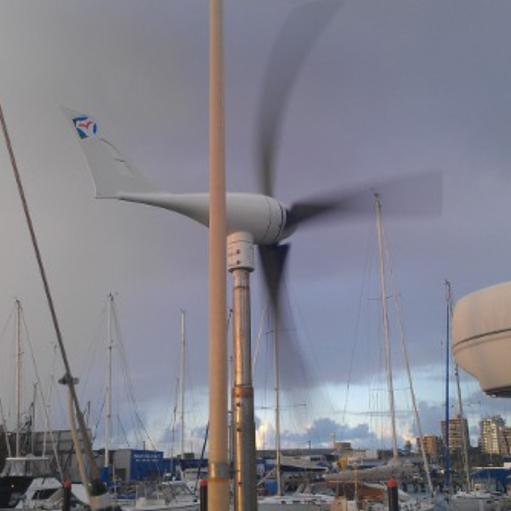 Wind turbine in operation at a marina, with blurred spinning blades and sailboats in the background.