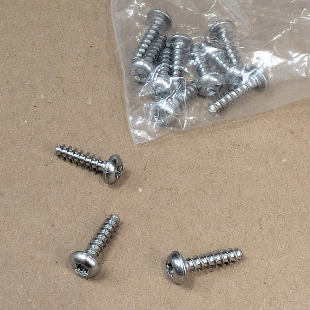 Several silver metal self-tapping screws with flat heads are shown on a brown cardboard surface, with a clear plastic bag containing more screws in the upper right.