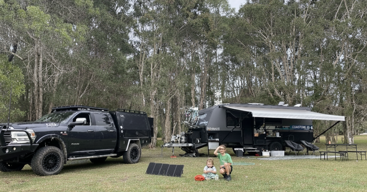 Off-grid campsite with a large black truck, trailer, two children, and portable solar panels in a grassy, wooded area.