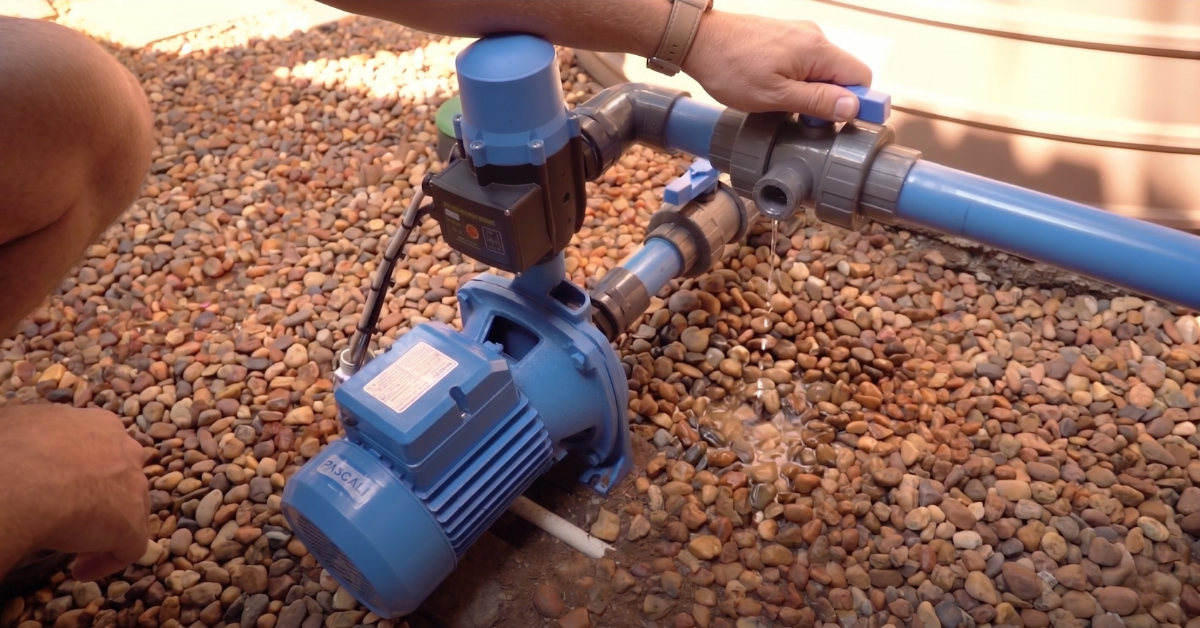 A person inspects a blue Pascali water pump and PVC pipes for leaks outdoors on a bed of small brown river stones.