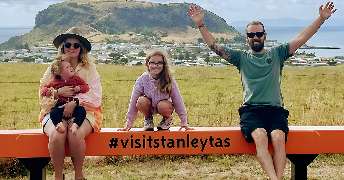 A family sitting in a scenic photo frame in Stanley, Tasmania, smiling with a picturesque backdrop and the hashtag #visitstanleytas.