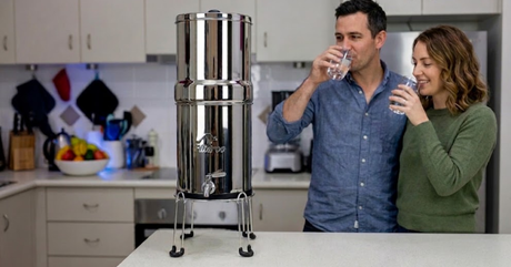 A couple drinks water from glasses next to a large stainless steel gravity water filter in a kitchen.