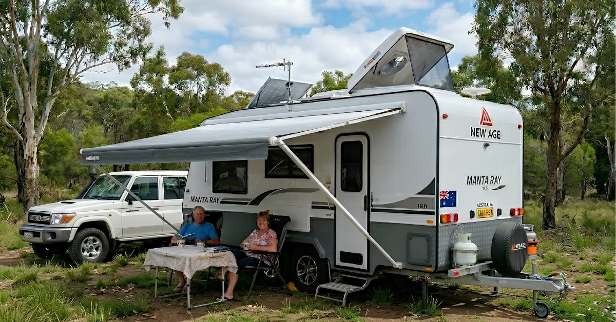 A couple sits under a caravan awning outdoors beside a 4WD, with a large roof hatch tilted open for ventilation.