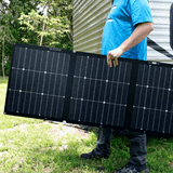 Person holding a large solar panel outdoors with trees and a trailer in the background