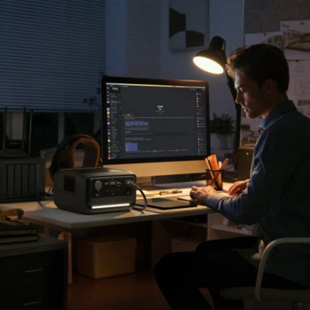 Man working at night with desktop powered by portable power station – A person focused on their work in a dimly lit room, using the power station to run a dual-monitor computer setup.