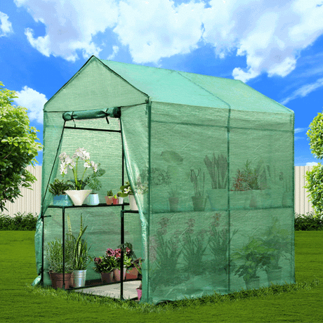 A small, walk-in greenhouse with a green, mesh fabric cover and an open zipped door is set on a lawn with various potted plants inside.