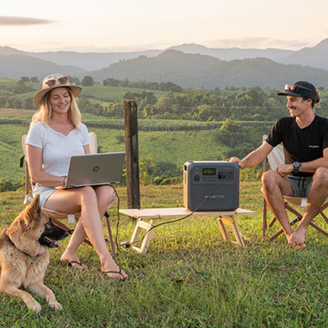 A lifestyle photo featuring two people outdoors using the Bluetti system to power a laptop, symbolising its practicality and versatility for outdoor and remote applications. A dog is also visible in the foreground.