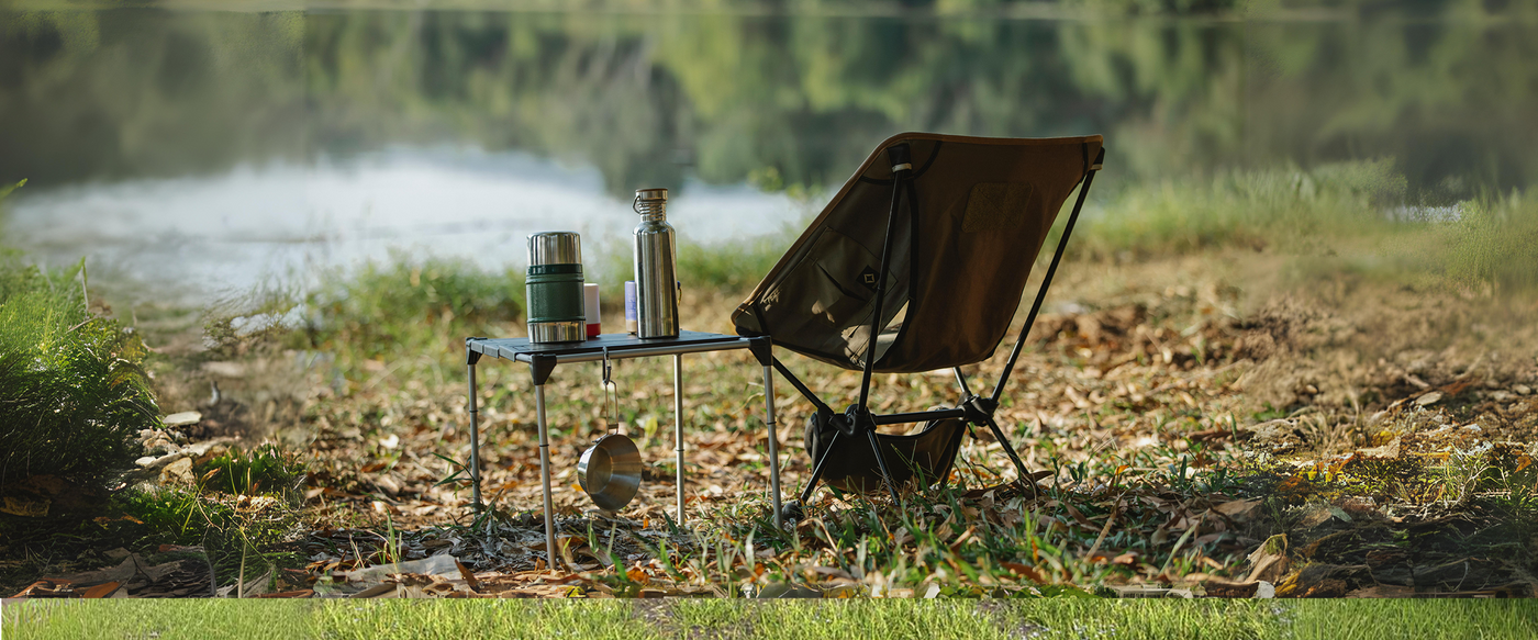 Outdoor camping setup by a serene lakeside with a brown folding chair and a small table holding a thermos, water bottle, and camping gear