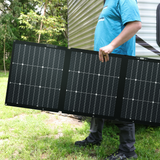 Person holding a large solar panel outdoors with trees and a trailer in the background