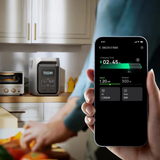 Person holding a smartphone displaying energy monitoring app in a kitchen.