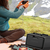 Person sitting outdoors with a phone and extra battery, surrounded by nature.