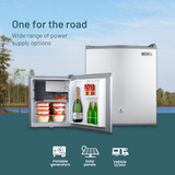 Portable refrigerator with open door showing drinks, set against a scenic background with trees and water.