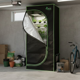 A black Green Fingers hydroponic grow tent sits in a garage, featuring reflective lining and internal plant shelving.