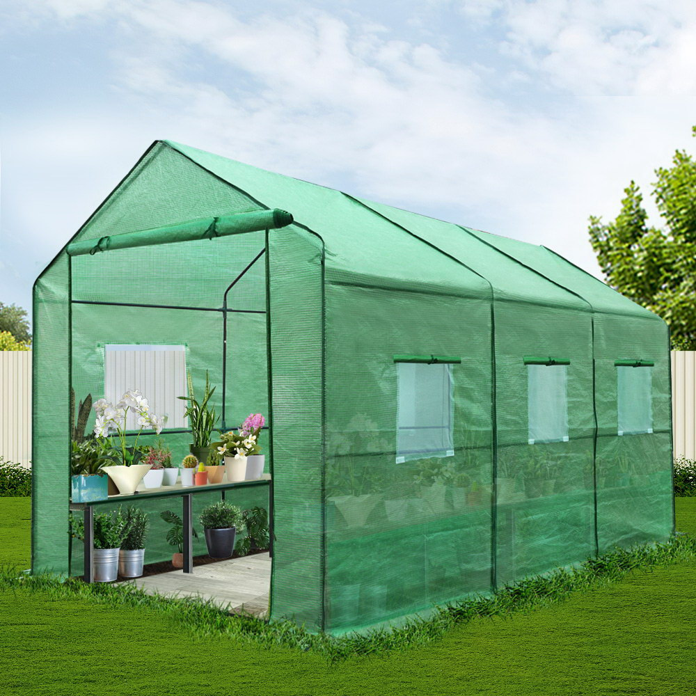 Green greenhouse with plants inside on a grassy area with trees in the background