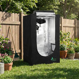 Black grow tent with transparent door on a grassy area with plants and a wooden fence in the background.