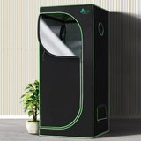 Black grow tent with green trim next to a potted plant on a wooden floor.
