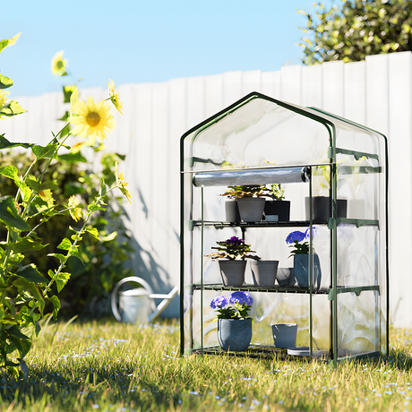 The mini greenhouse in an outdoor garden setting, filled with small potted plants. The greenhouse stands on grass with sunflowers nearby, adding a touch of nature to the scene