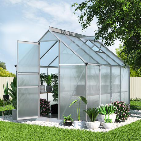 An outdoor setting featuring the aluminium polycarbonate greenhouse with plants and flowers inside and surrounding the structure, showcasing the greenhouse in a lush garden environment