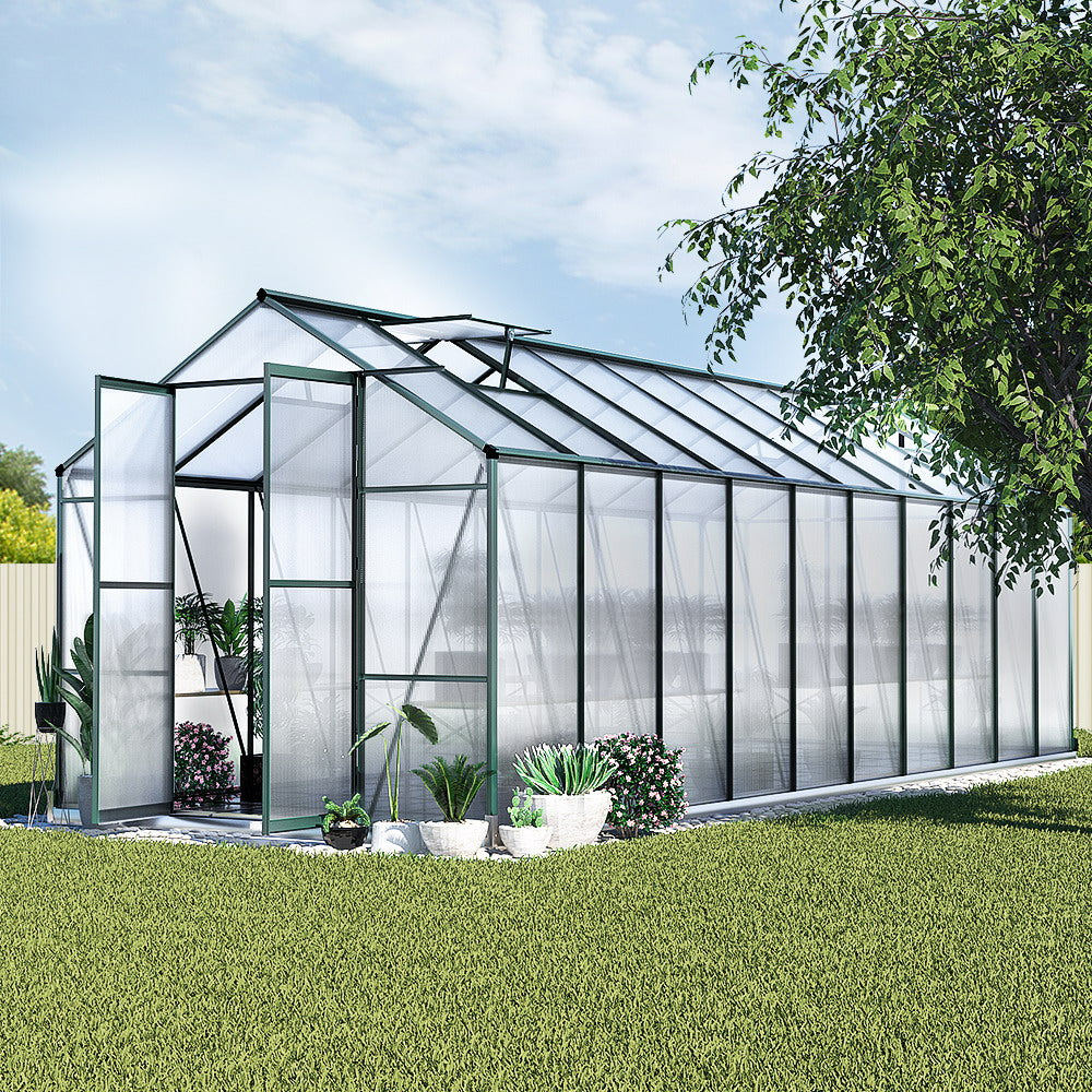 A spacious aluminium polycarbonate walk-in greenhouse in a garden, surrounded by potted plants, under a clear sky.