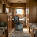 The toilet is placed in the center aisle of a vehicle with a wood-paneled interior, likely a vintage caravan or RV. Sunlight is coming through a window in the background.
