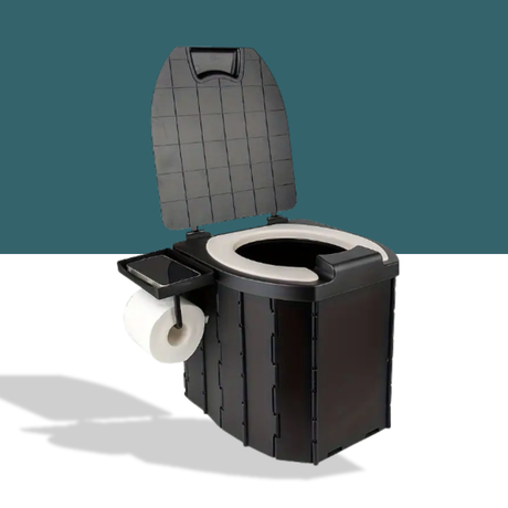 A white portable toilet is placed outdoors on a forest floor, positioned in front of a blue camping tent.