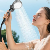 Person using a handheld shower head with water spraying onto their face against a clear sky.