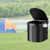 The white portable toilet is placed outdoors on a forest floor, positioned in front of a blue camping tent, showcasing its use in a rugged outdoor setting.