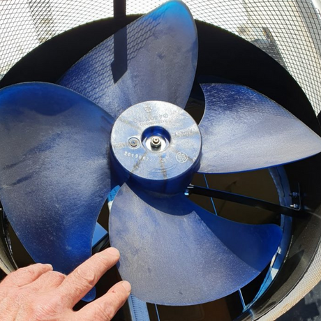 Close-up of a blue metal fan blade inside a roof ventilator, with a hand pointing for scale.