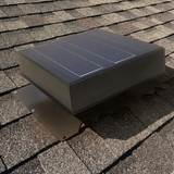 A black solar roof vent with a flashing base is mounted on grey asphalt shingles, casting a realistic shadow.