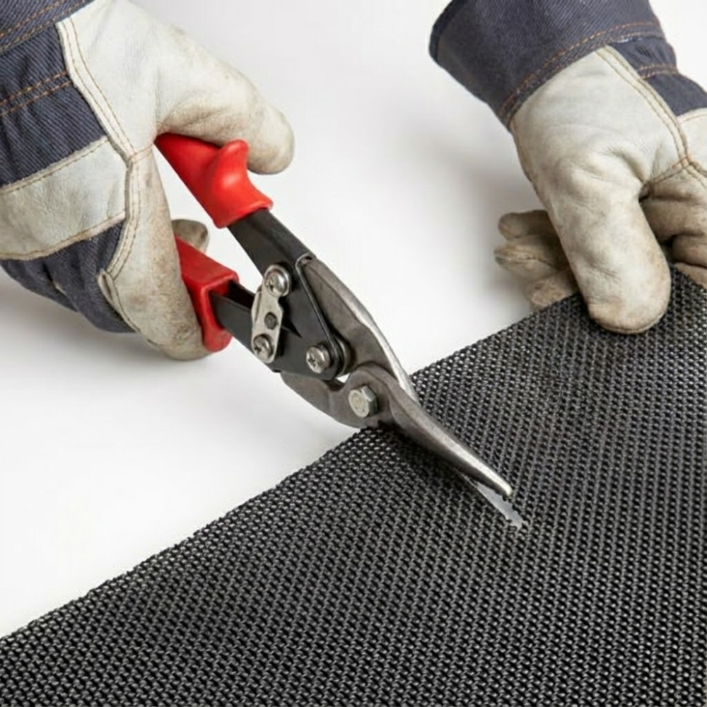 Gloved hands use red-handled tin snips to manually cut through the durable black metal mesh screen for a custom fit.
