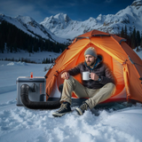 Person sitting in an orange tent in a snowy mountain landscape with a cooler and fan nearby.
